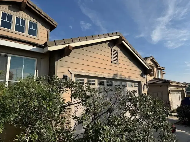 Suburban house exterior with tan siding, white trim, and bushes in front under a blue sky