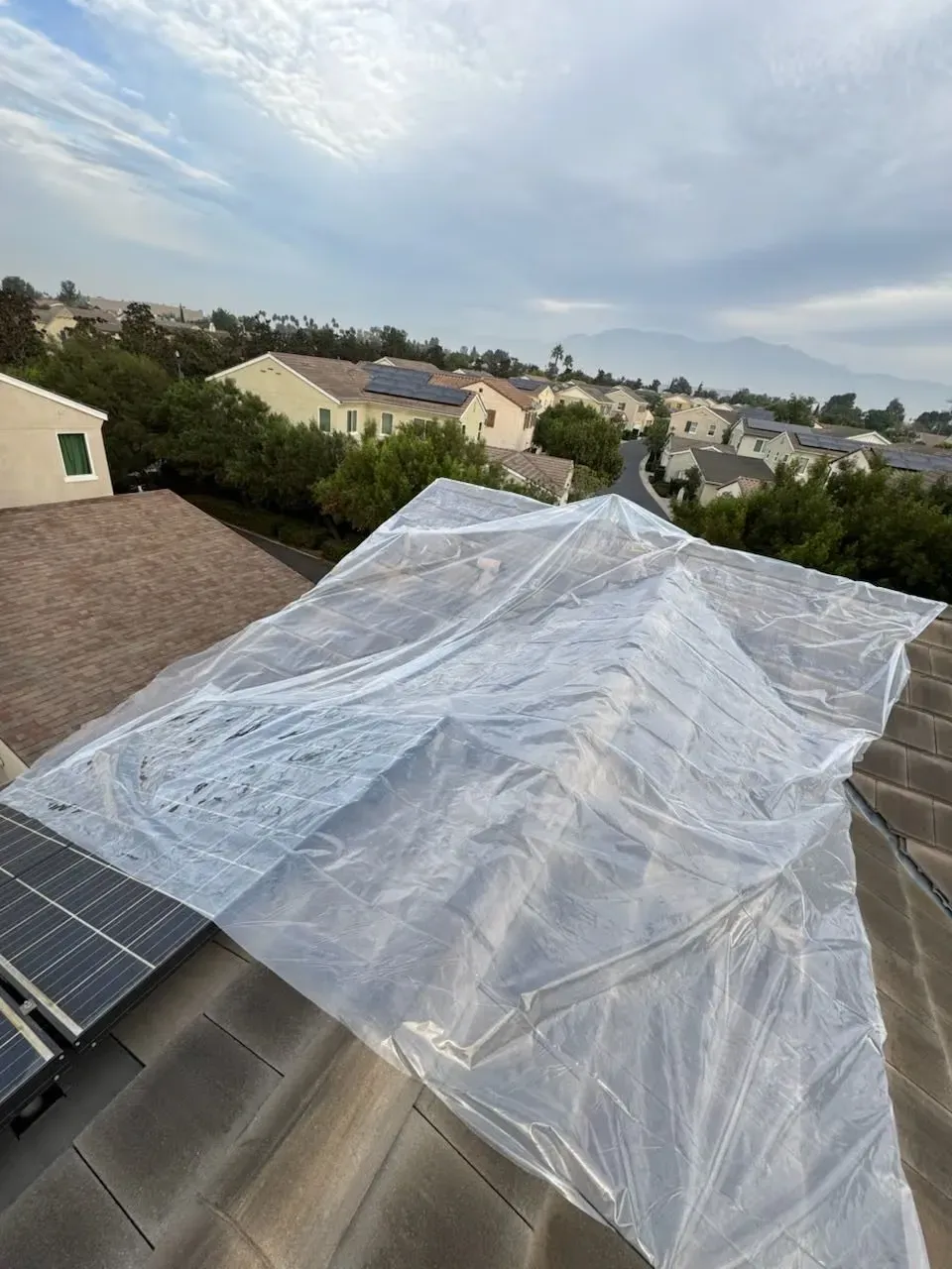 Clear plastic sheet draped over a rooftop near solar panels under a cloudy sky.