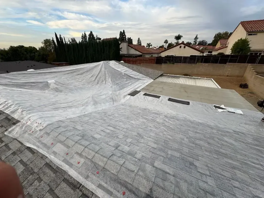 Partially waterproofed residential roof with white tarp under cloudy sky