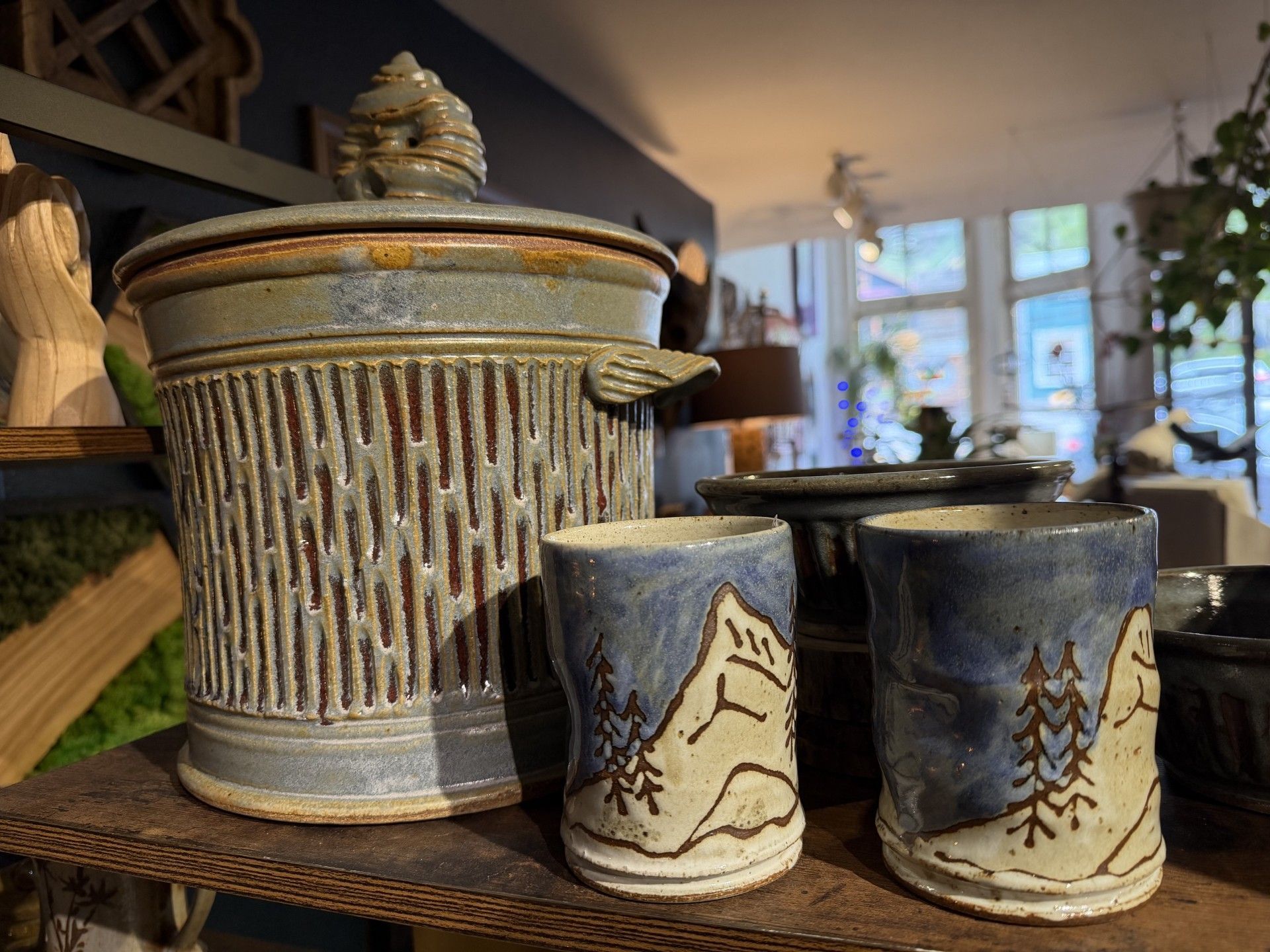 Three ceramic containers on a table, including a ribbed lidded jar and two painted mugs.