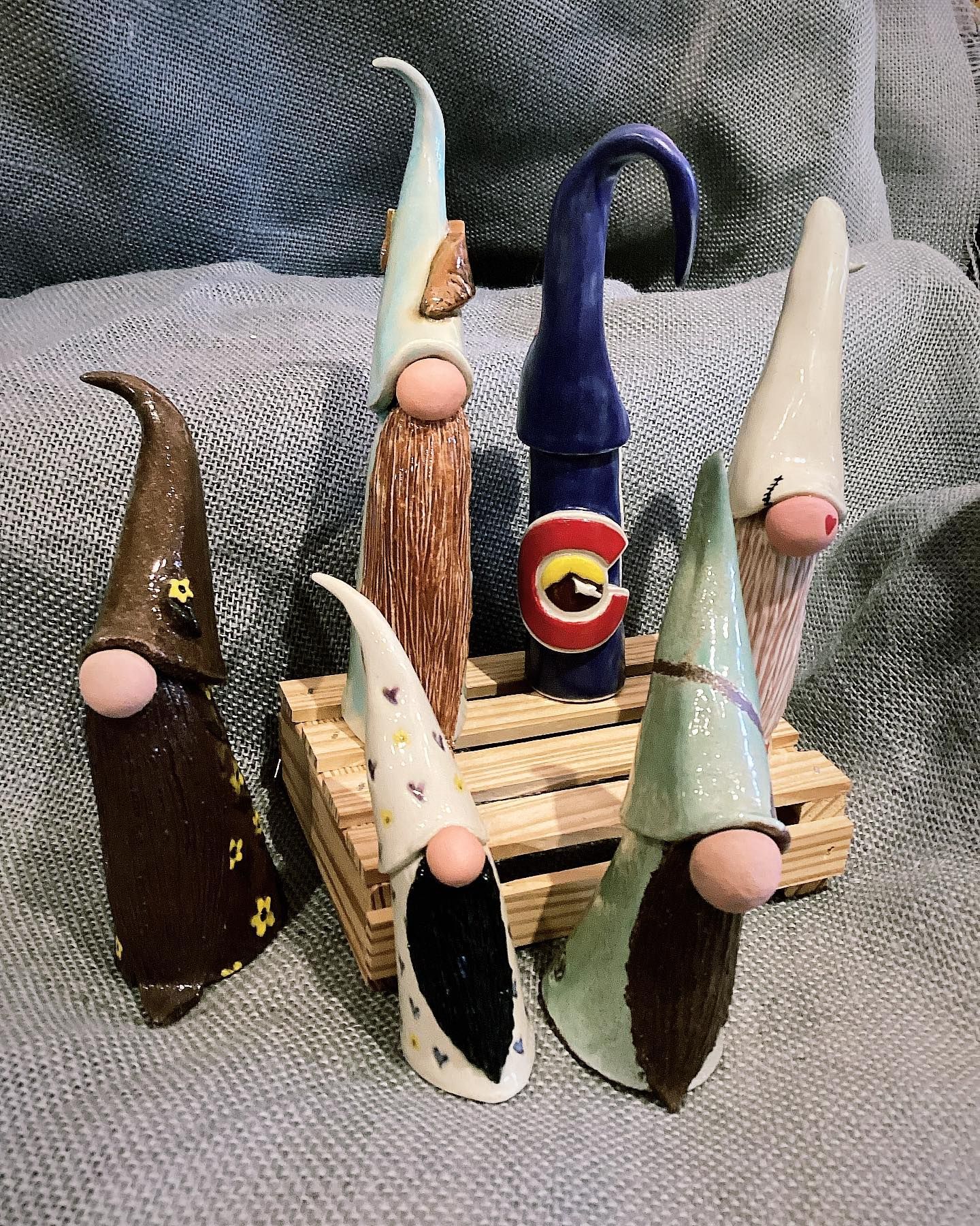 Seven colorful handmade cone-shaped figurines on a small wooden pallet, photographed against gray fabric.