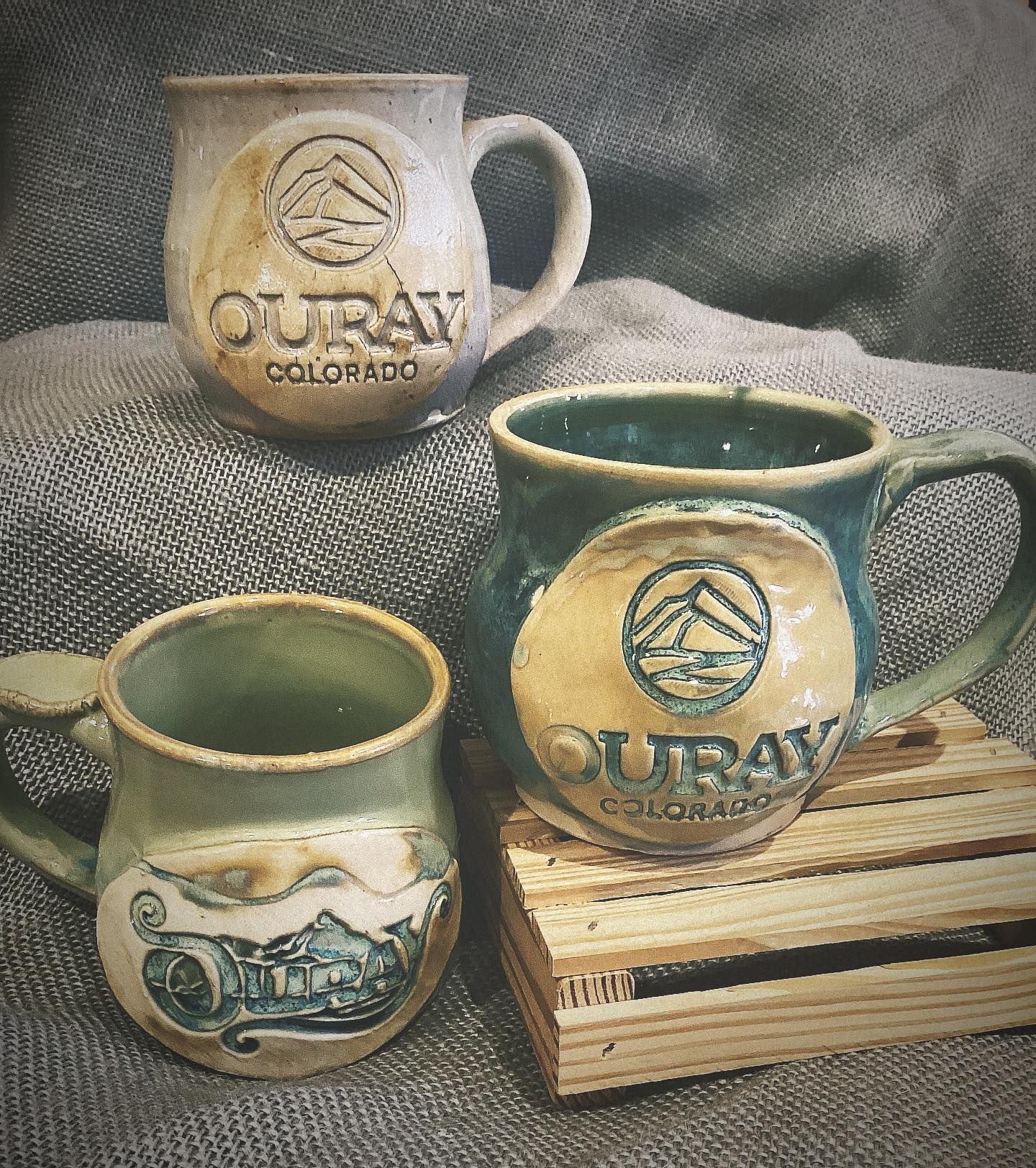 Three rustic ceramic mugs in cream, blue, and green on a gray cloth, one on a wooden crate.