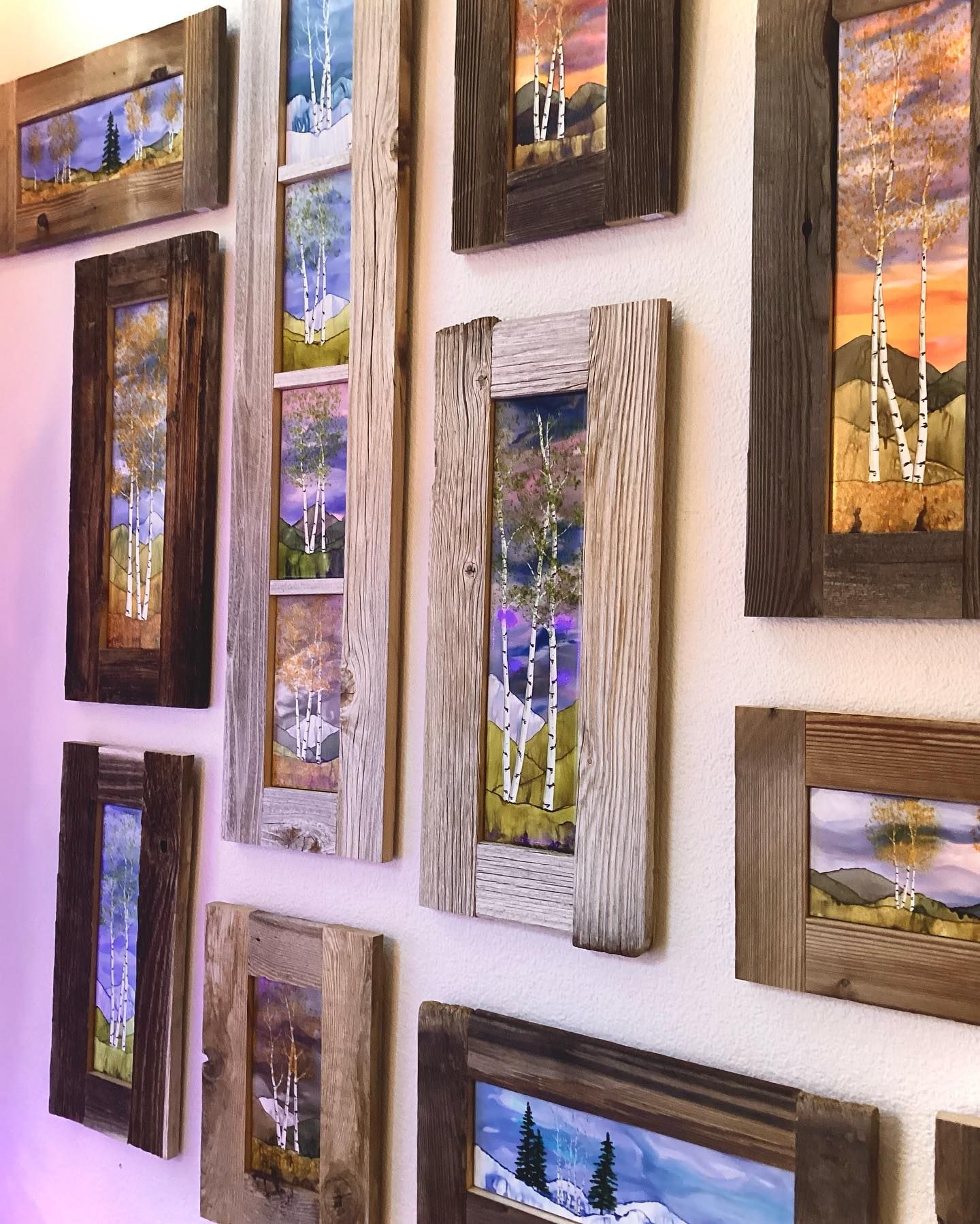 Assorted framed landscape paintings arranged on a white wall, featuring colorful sky, trees, and water scenes