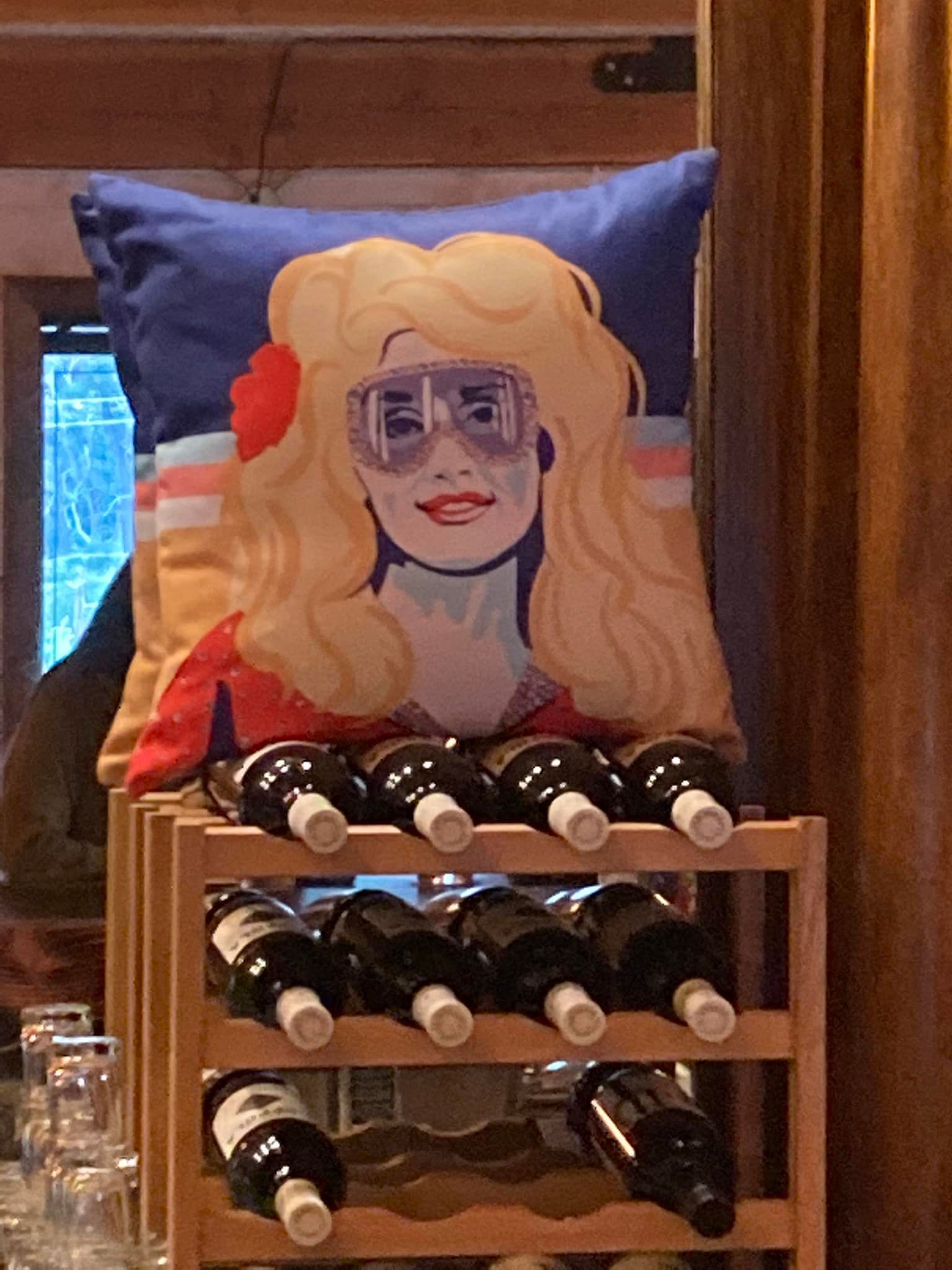 Decorative pillow with a blonde cartoon face sits on a wooden wine rack filled with bottles.