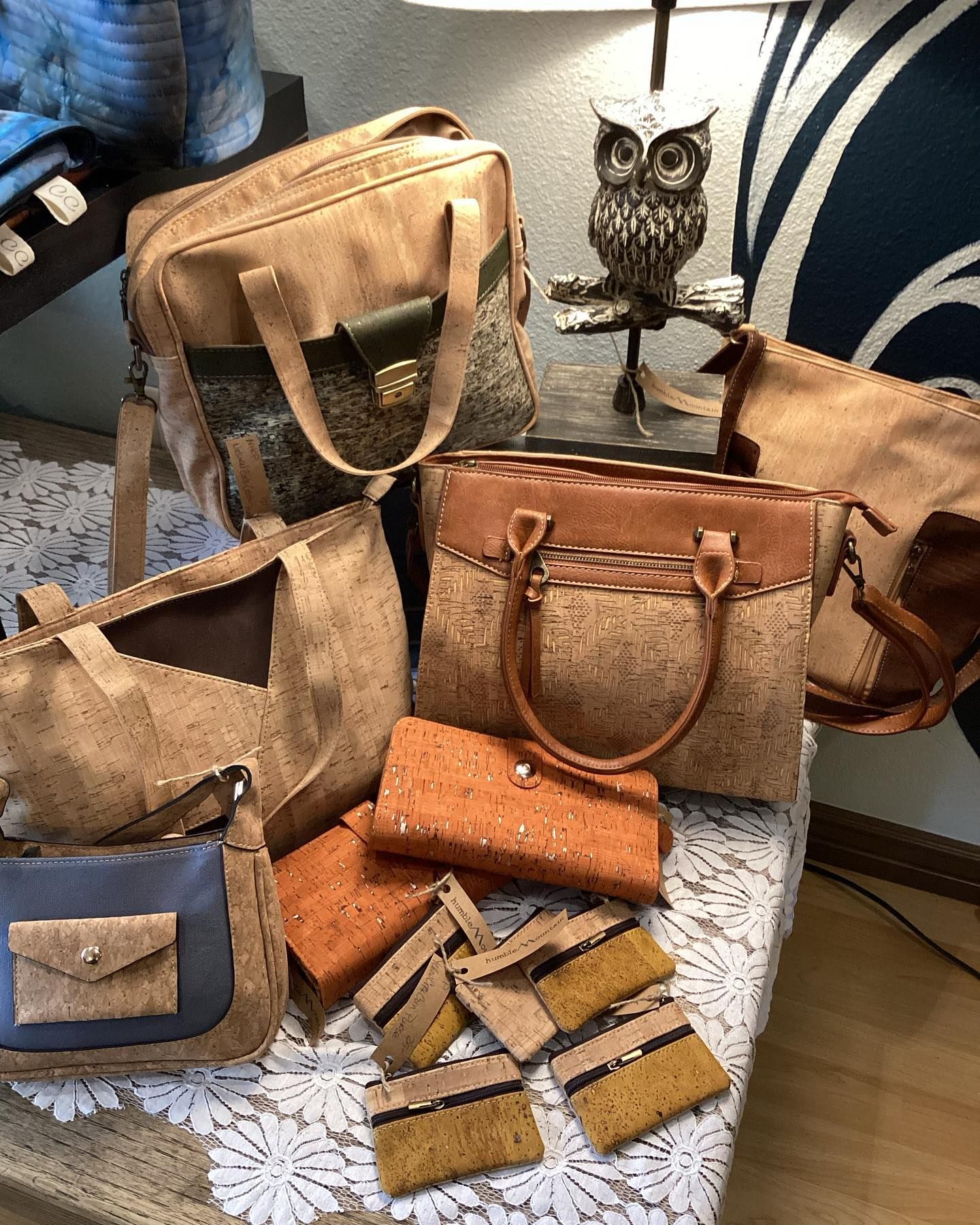 Assorted tan and brown handmade leather bags and wallets displayed on a table, with an owl figurine behind them.