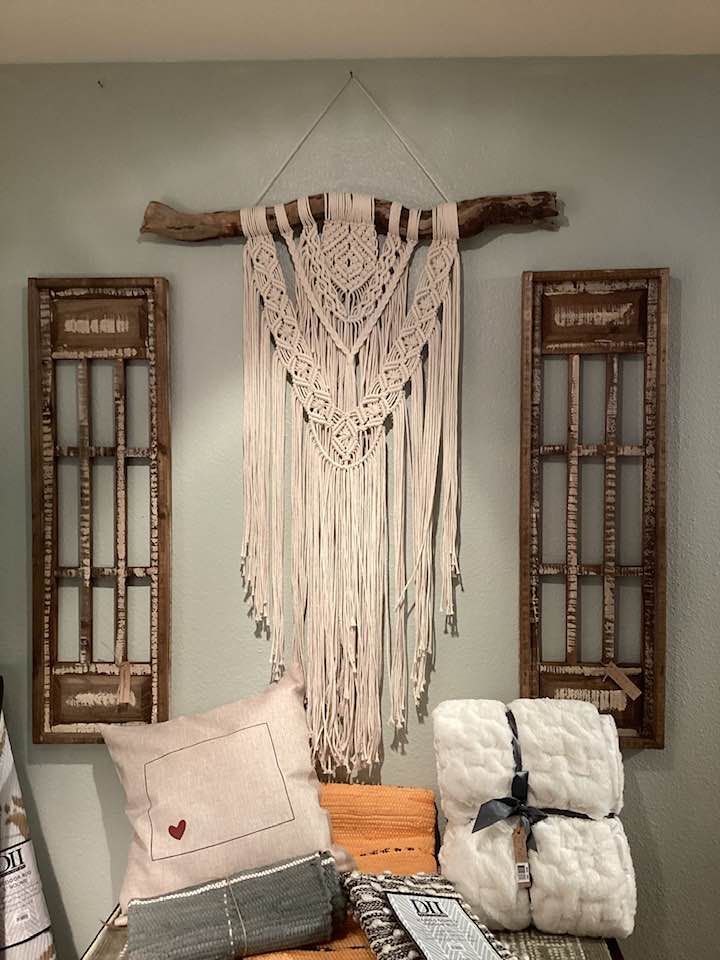 Macramé wall hanging on a wood branch above pillows and a bench against a pale wall