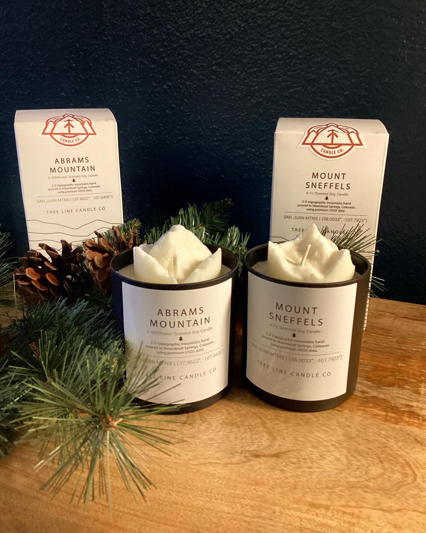 Two scented candles with pine and pinecones on a wooden surface against a dark background