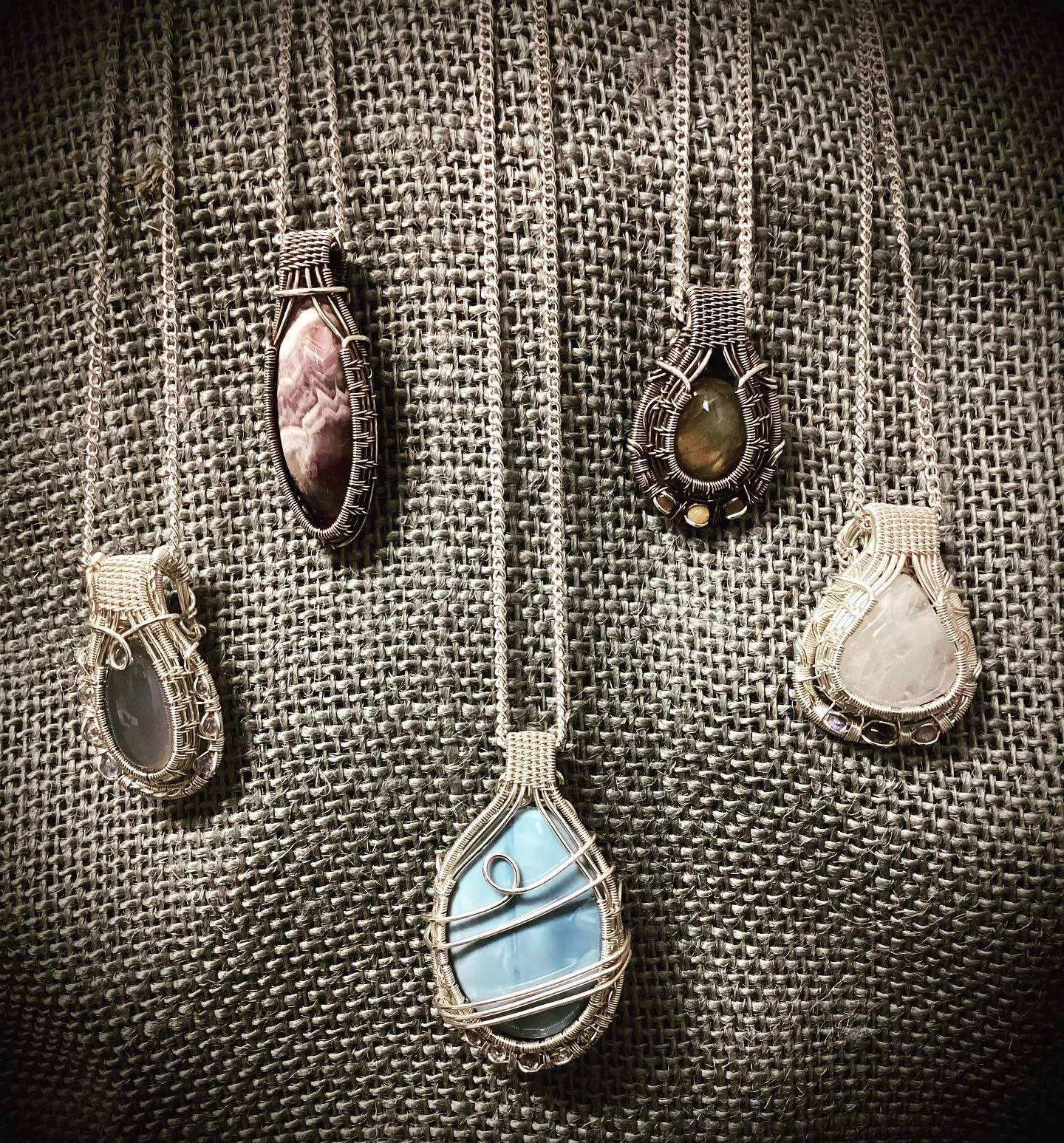Five teardrop pendant necklaces on textured fabric, with stones in blue, white, amber, and mauve.