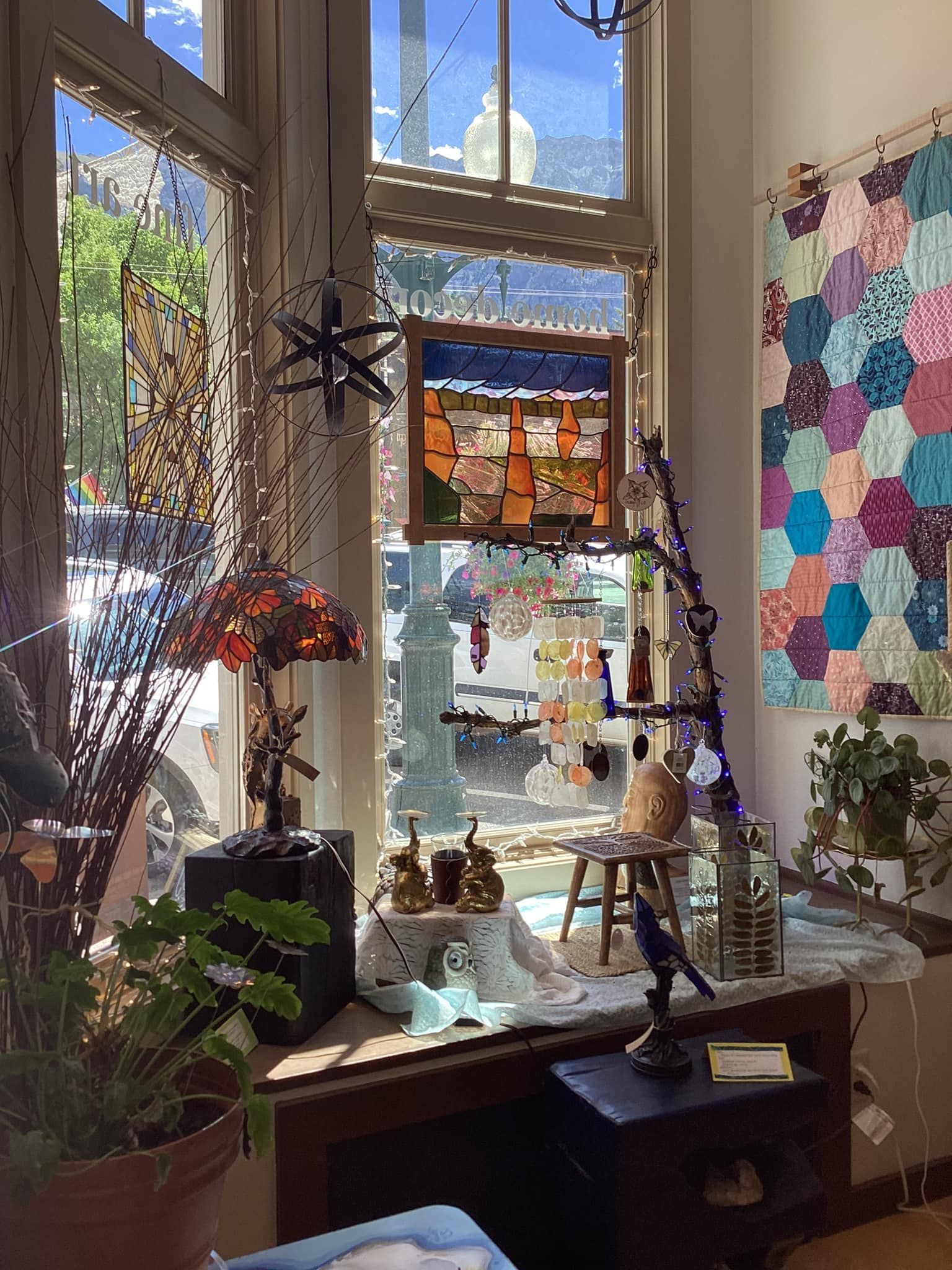 Cozy sunlit room with a large window, plants, hanging ornaments, and colorful quilted wall art
