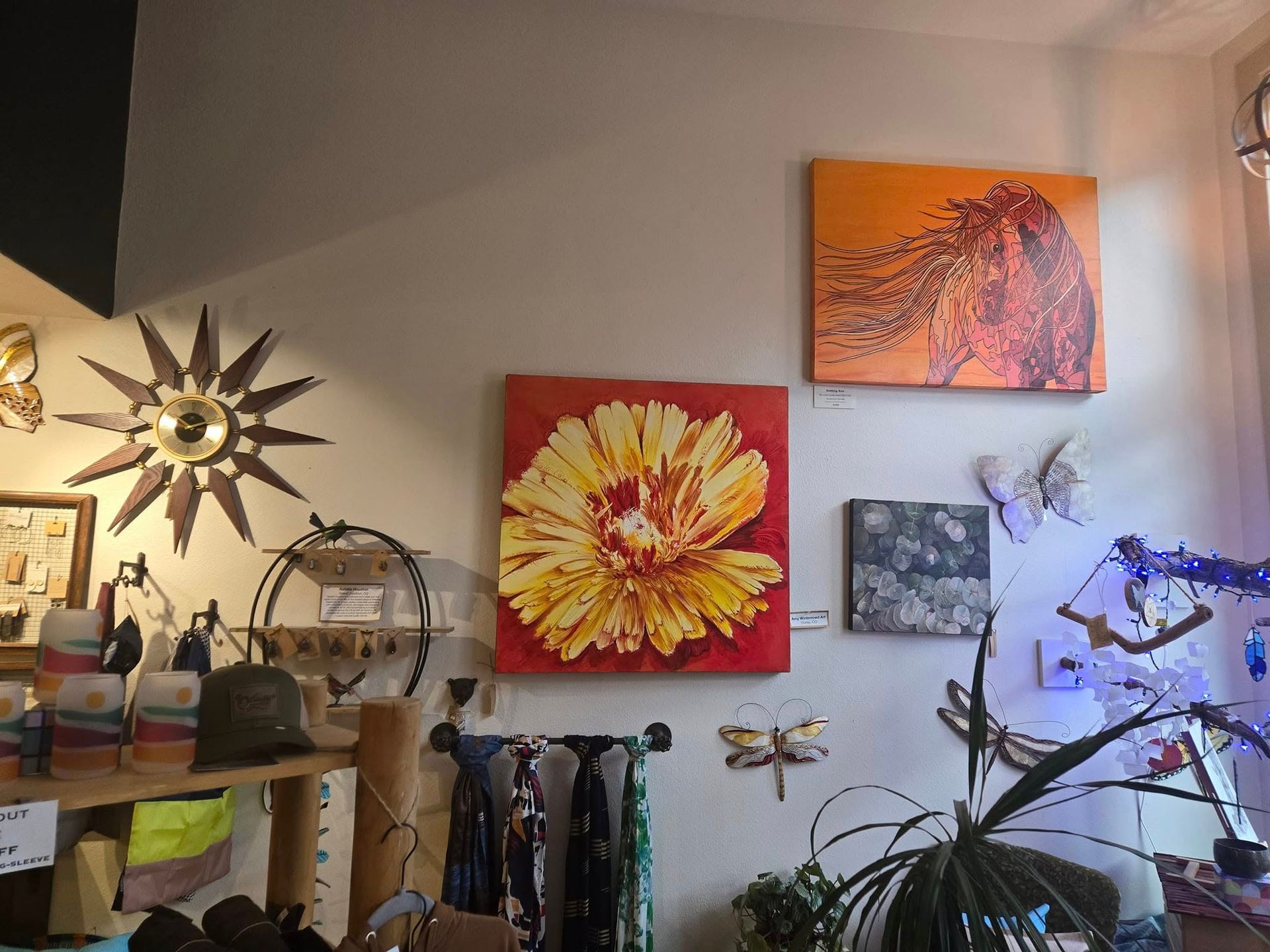 Wall with colorful abstract floral paintings, a sunburst mirror, shelves, and a plant lit by blue string lights