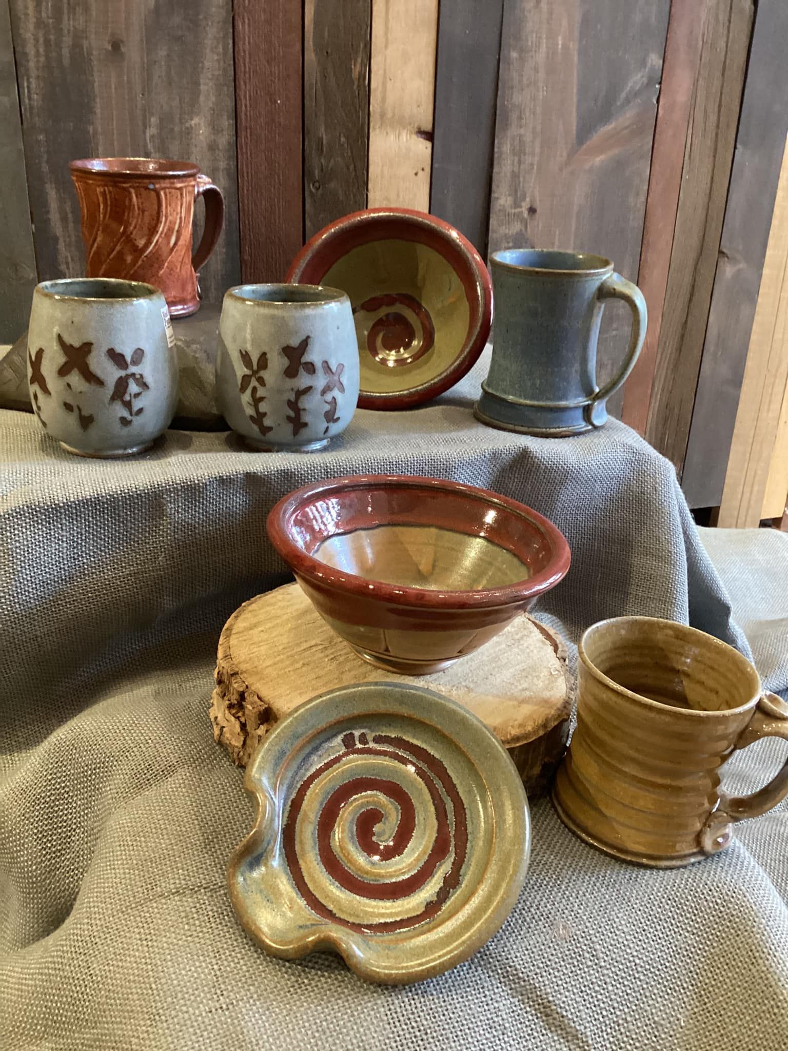 Assorted handmade ceramic bowls, cups, and mugs arranged on woven mats against a rustic wooden backdrop