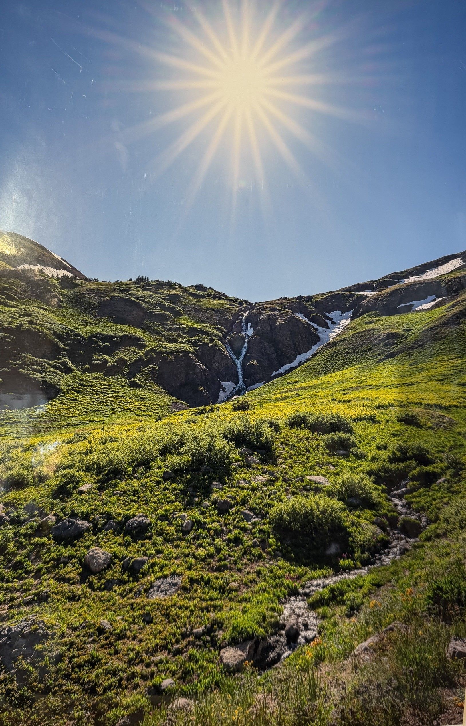 Sunny mountain meadow with green hills, rocky slopes, and a blue sky with bright sun rays