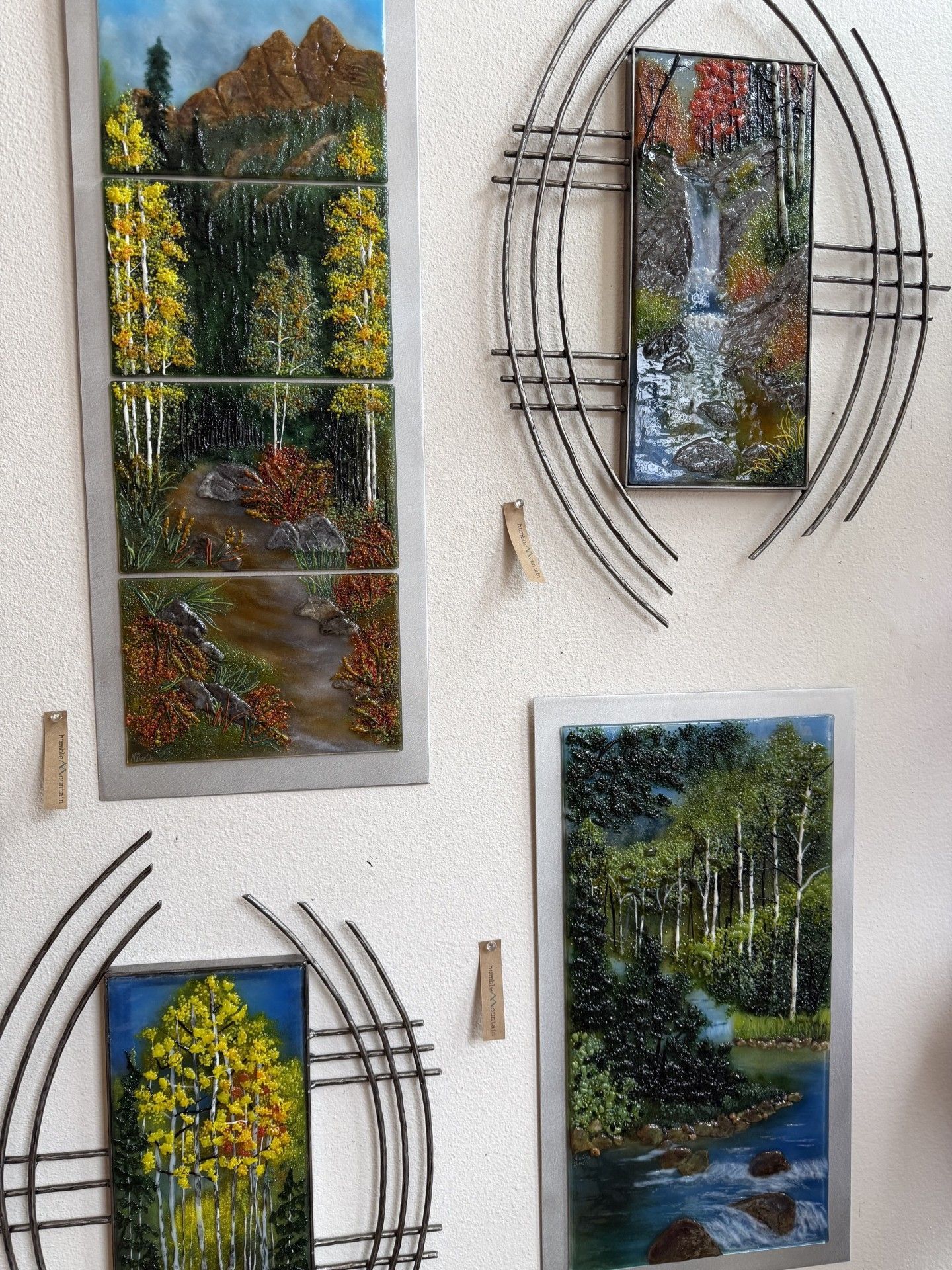 Four small framed nature paintings mounted on a white wall, including a forest scene and autumn trees