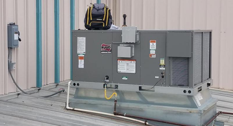 A rooftop HVAC unit with a technician's backpack sitting on top, positioned against a metal wall with a power box.