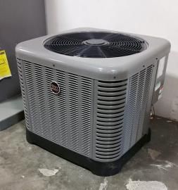 A gray, square Rheem HVAC outdoor condenser unit sitting on a concrete floor.