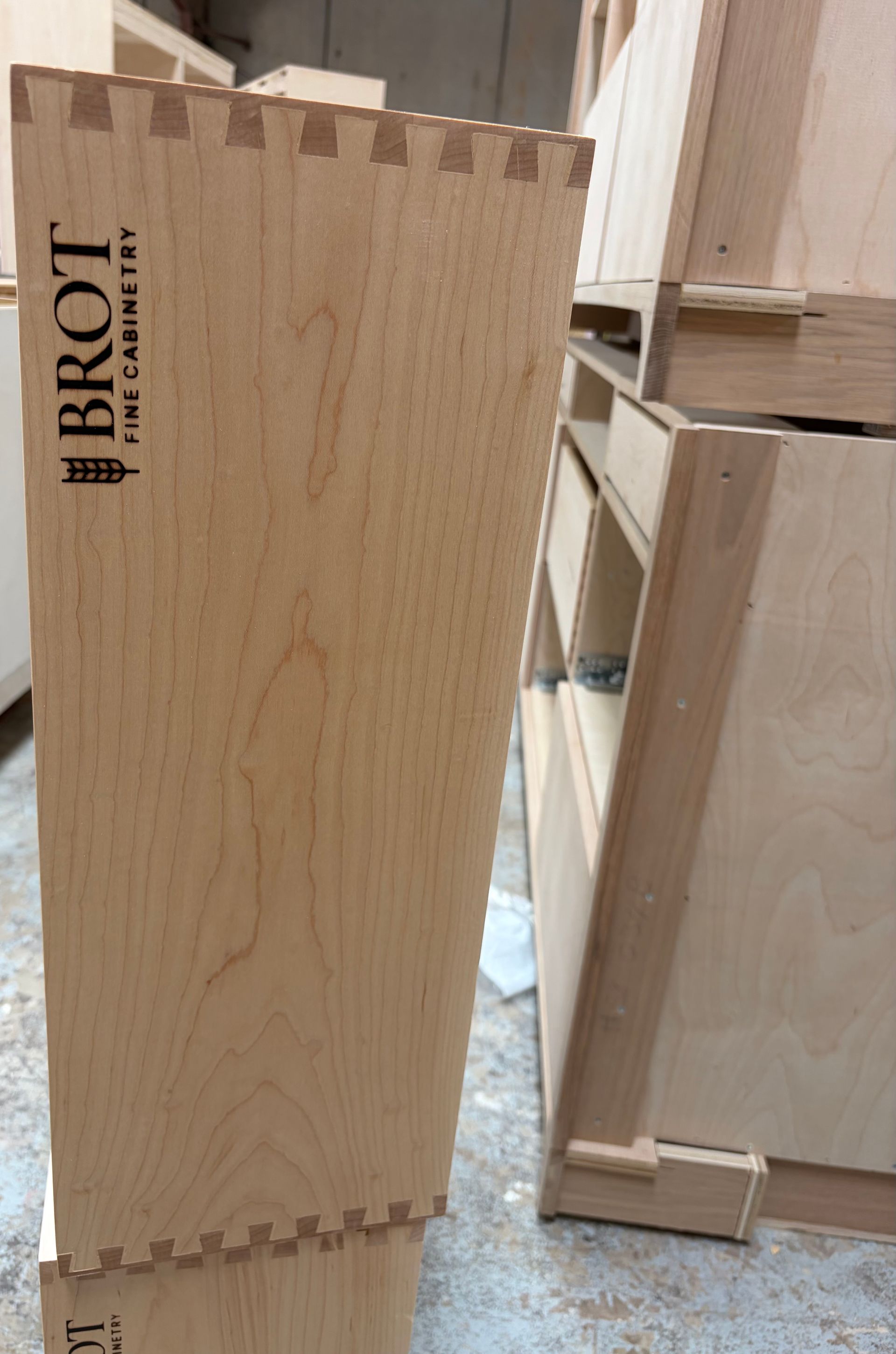 Wooden crate panel with dovetail joints and a black logo, beside other wooden boxes on a workshop floor