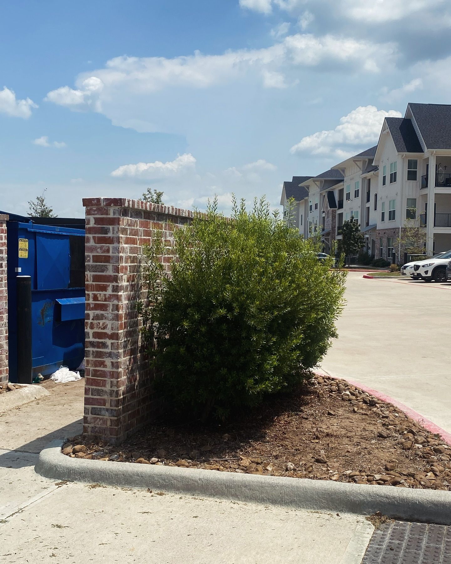 A blue dumpster behind a brick wall with a shrub in front, near an apartment building and parking lot.