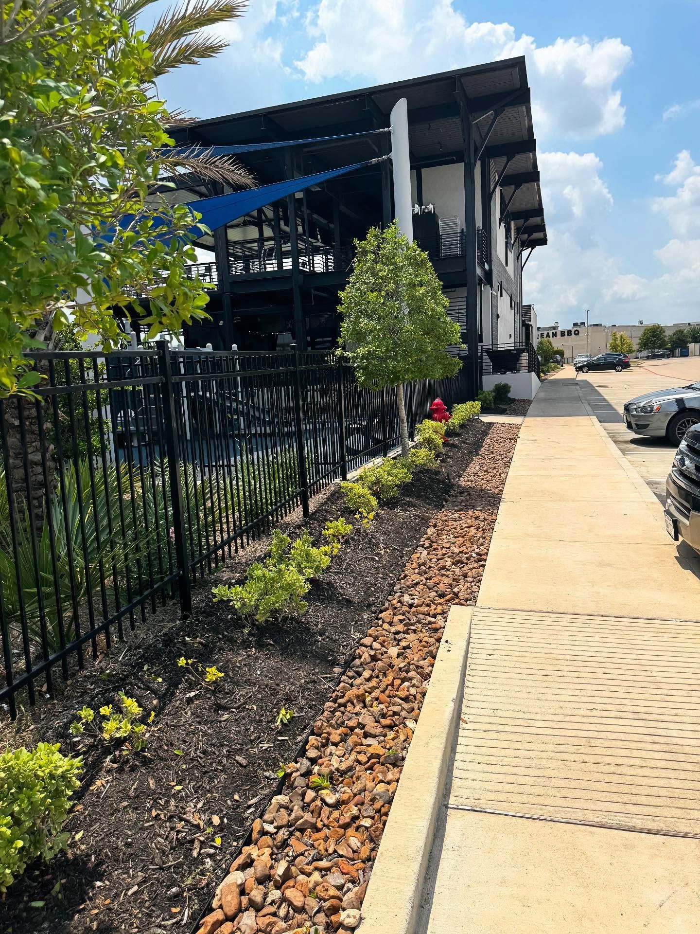 Sidewalk next to a building with black fence and landscaped bed with rocks and plants.