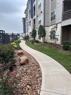 Curved concrete walkway beside apartment building with landscaping, rocks, and pink flowers