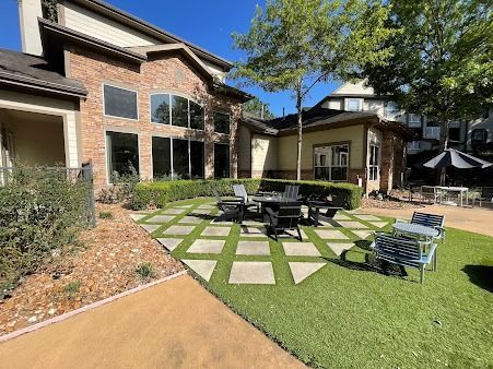 Outdoor seating area with tables and chairs in front of a glass building. Trees and green structures are visible.