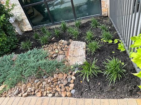 Small landscaped garden bed with rocks, dark mulch, and green ornamental plants beside a house and fence