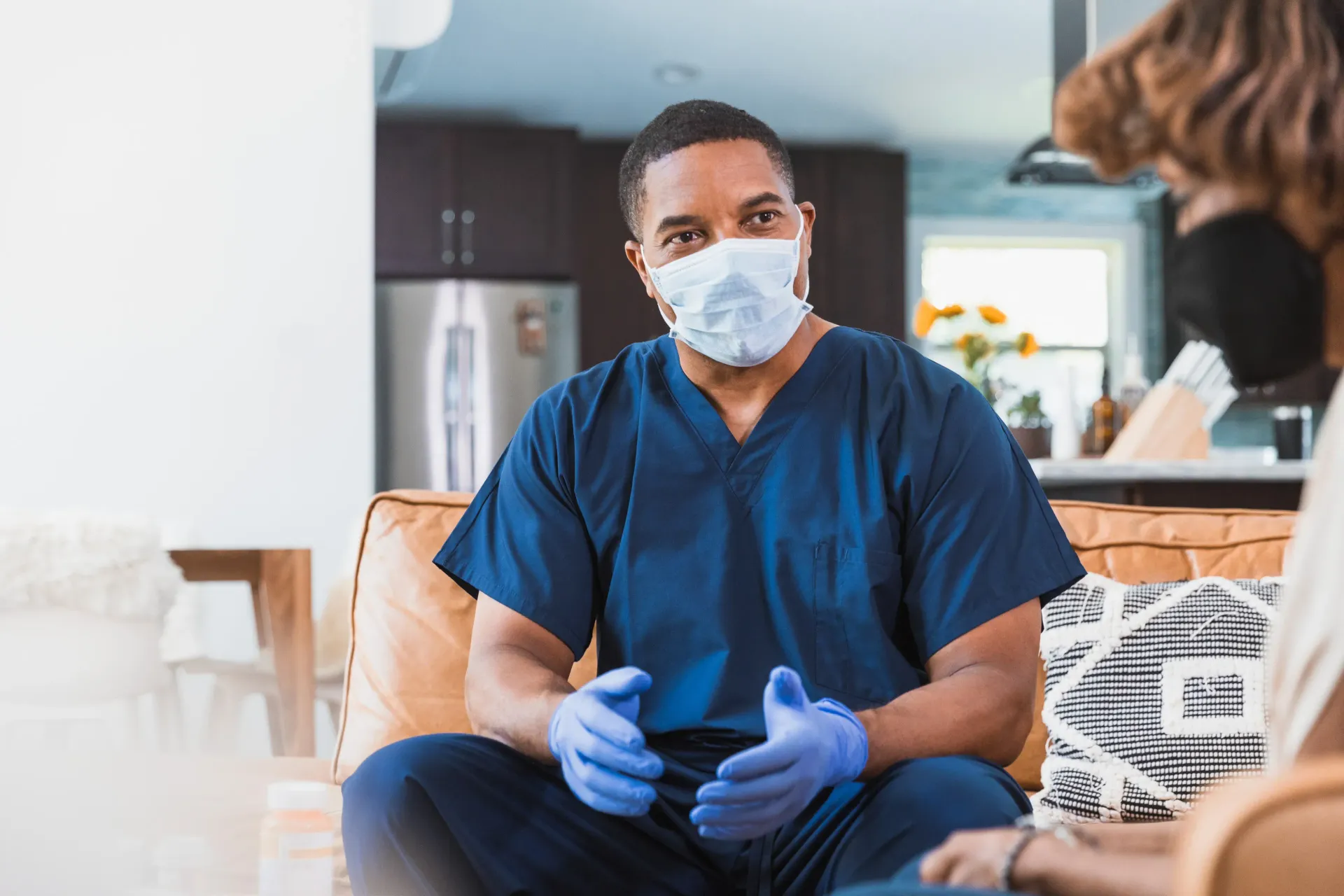 A nurse wearing a mask and gloves is sitting on a couch talking to a patient.