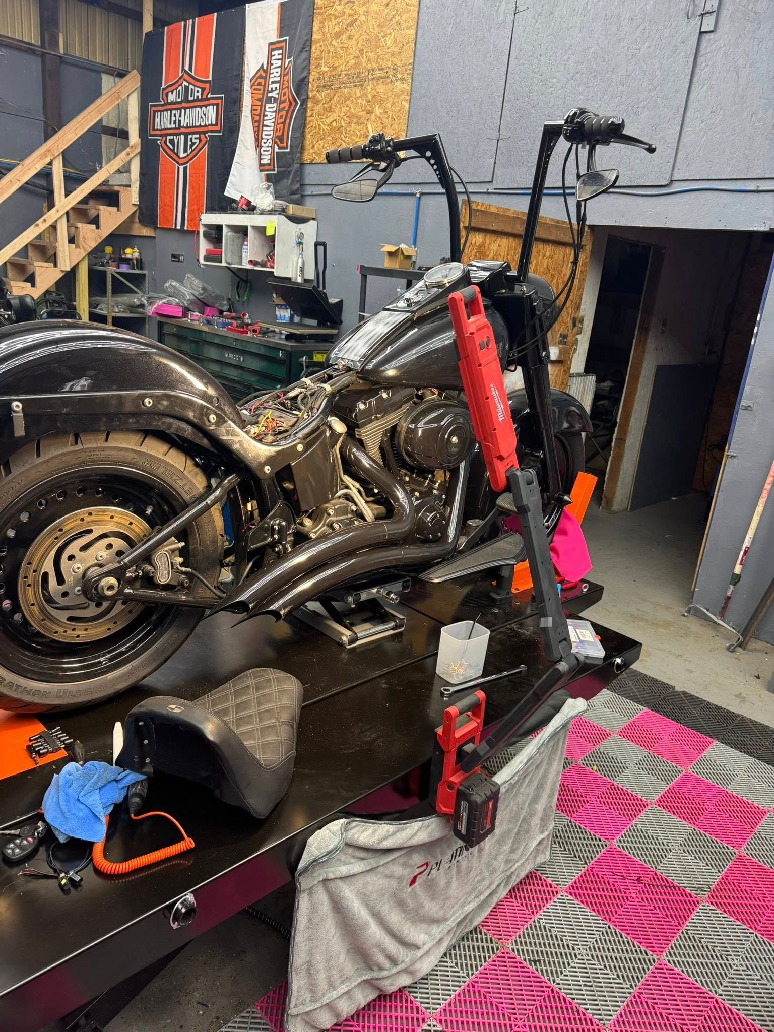 Black Harley Davidson motorcycle on a lift, undergoing repairs in a garage. Pink and black blanket visible.