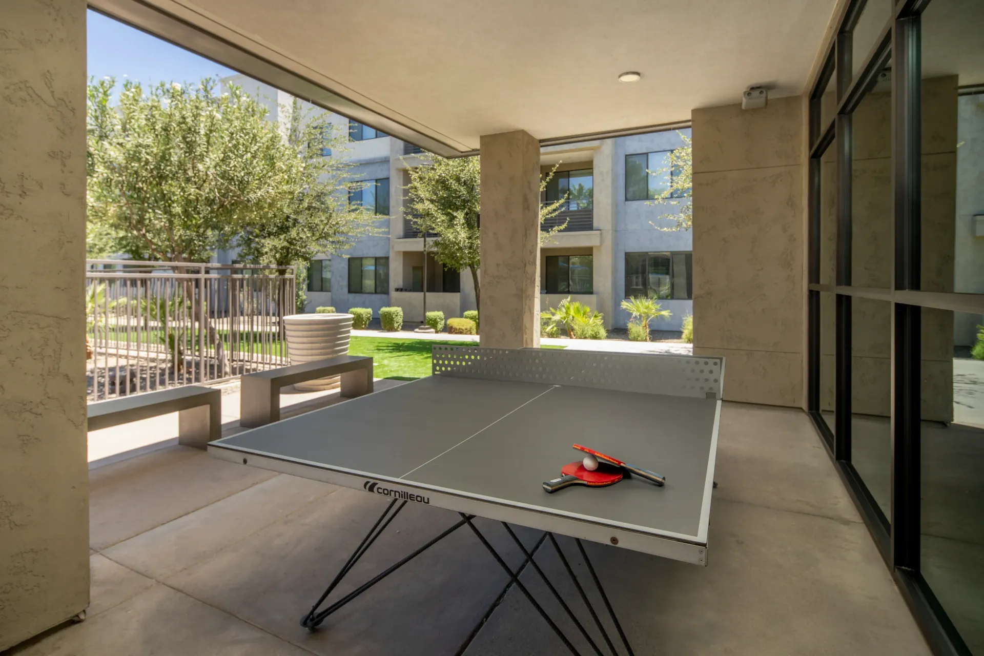 Ping pong table with paddles and ball on an outdoor patio with benches and courtyard view.