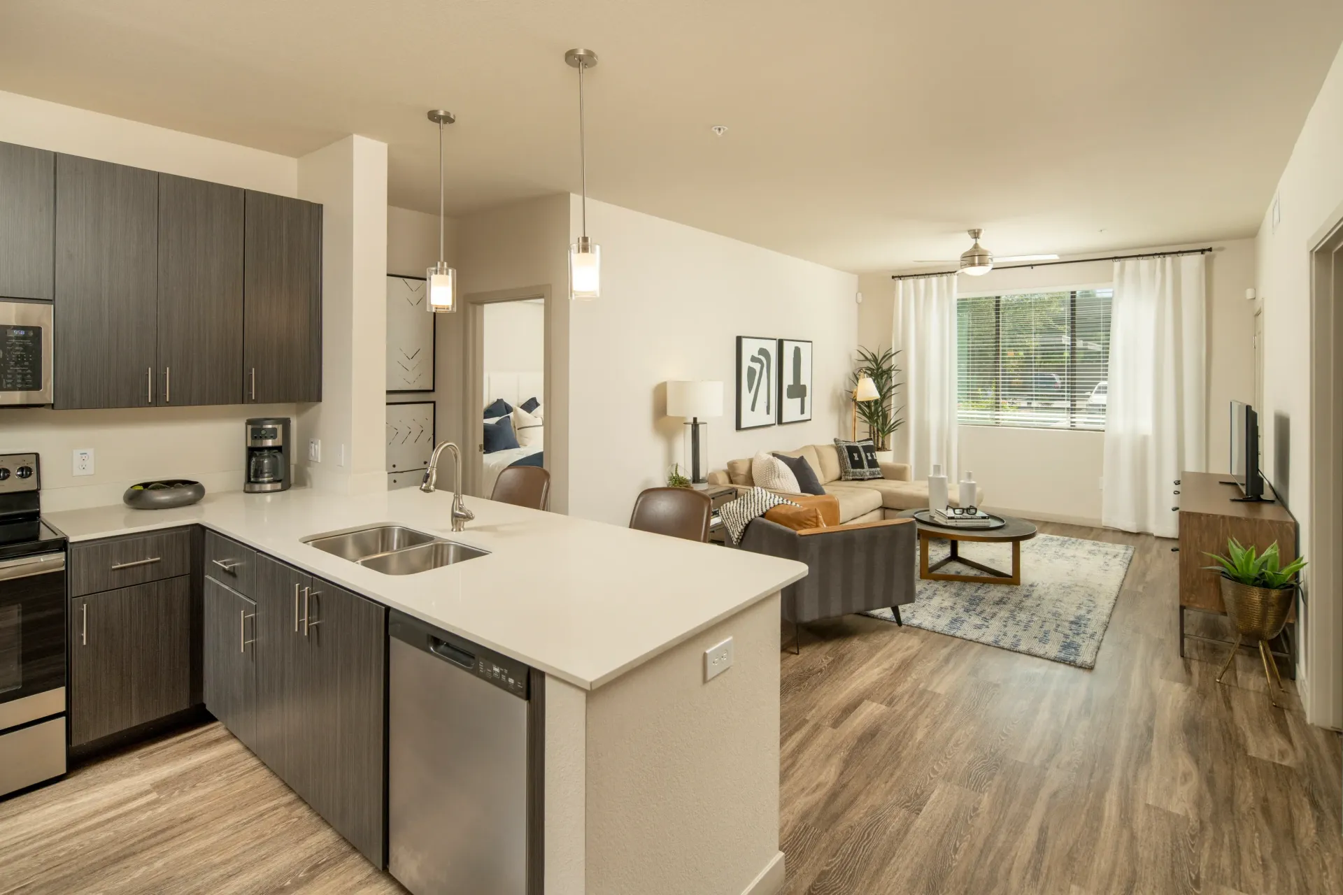 Modern apartment kitchen with stainless steel appliances and island, flowing into a living room with sectional couch and large window at Parc South Mountain in Phoenix, AZ.