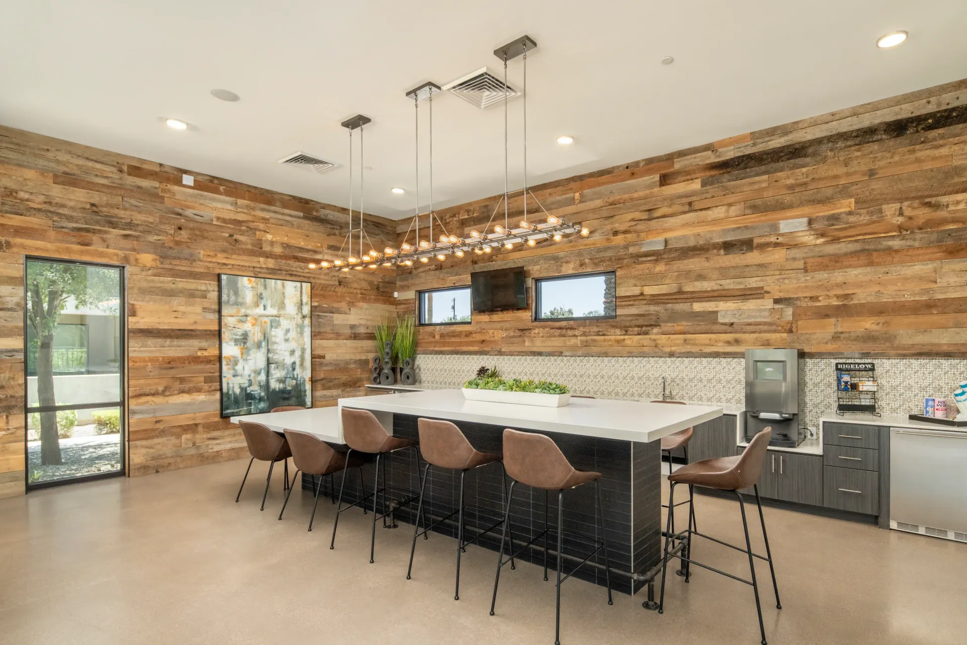 Modern communal lounge area with a long island bar, comfortable seating, and rustic wood accent wall at Parc South Mountain in Phoenix, AZ.