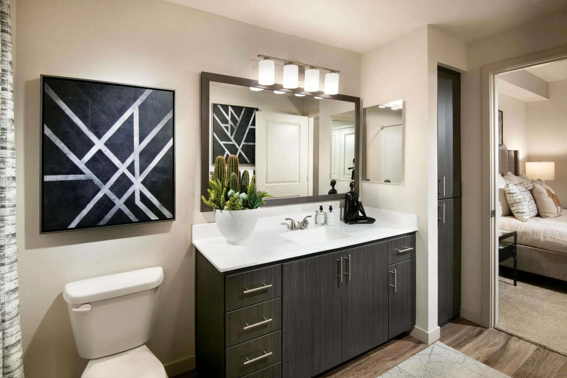 Modern bathroom vanity with dark wood cabinets, white countertop, large mirror, and contemporary lighting. A toilet is to the left, and a bedroom is visible through an open doorway. at Parc South Mountain in Phoenix, AZ.