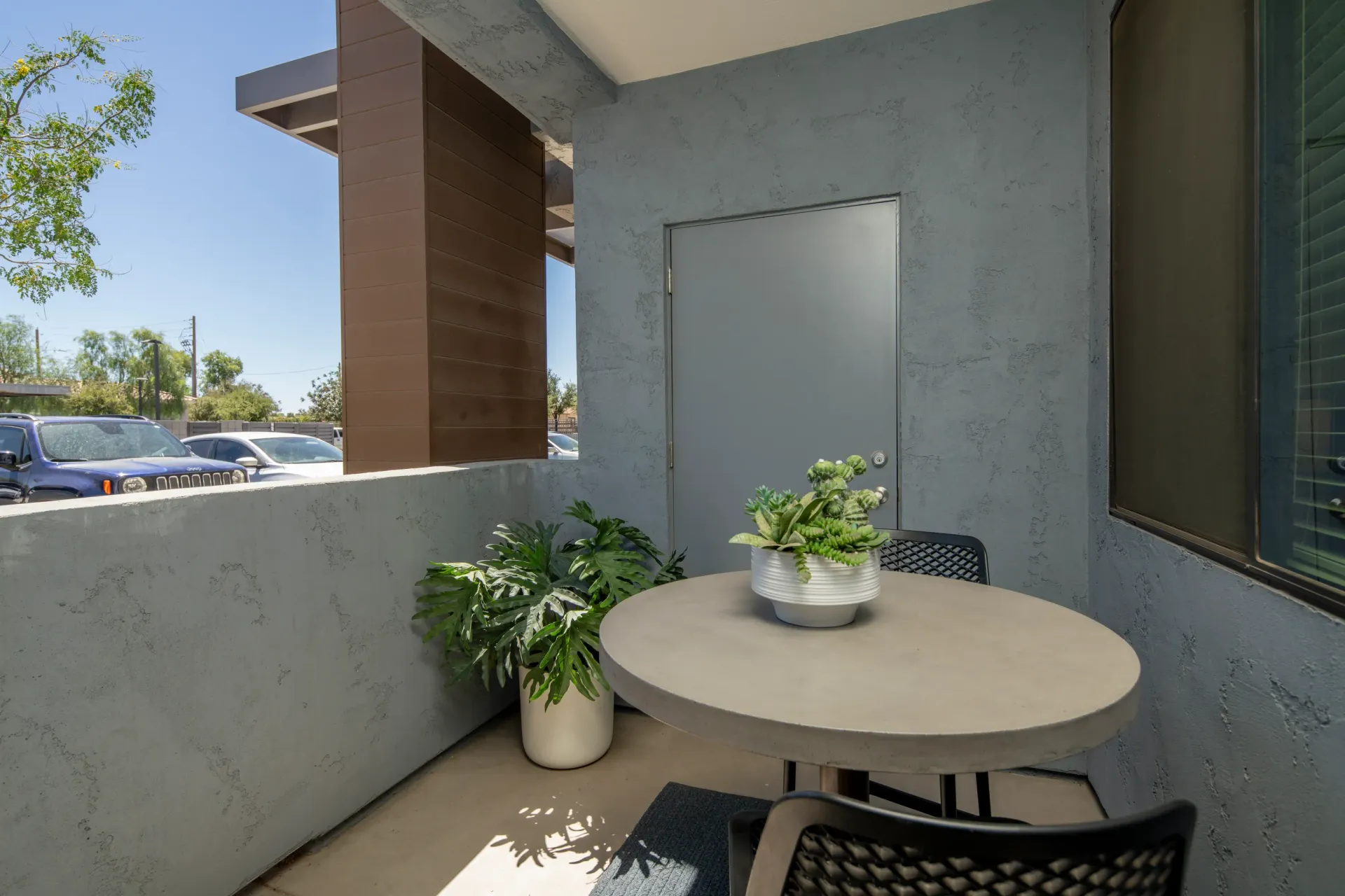 A small balcony with a round table and two chairs, decorated with potted plants and a grey door at Parc South Mountain in Phoenix, AZ.