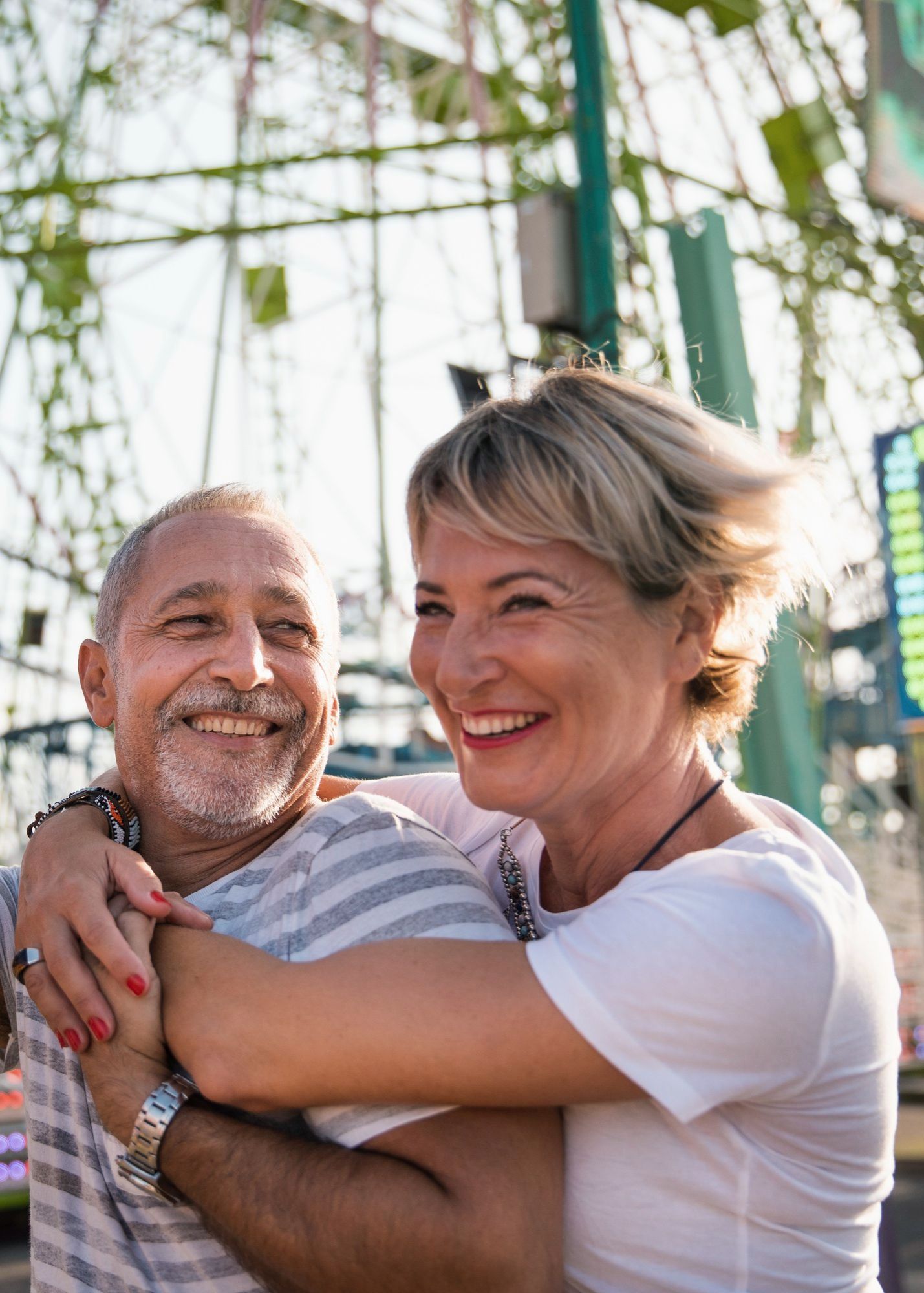 Mature couple embracing, smiling broadly. Woman with short blonde hair hugs man in striped shirt at a carnival