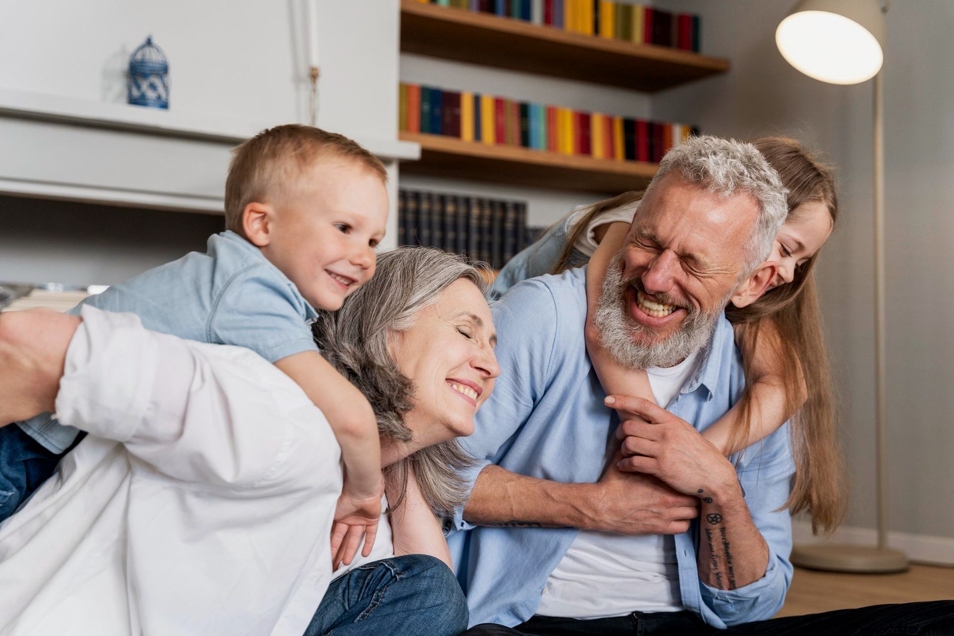 Grandparents laughing while being embraced by their grandchildren indoors; bookshelf in background.