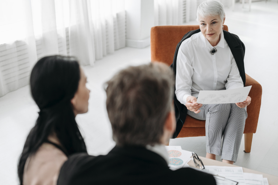 Three people in a bright office discussing paperwork, with a seated woman speaking and holding a document.