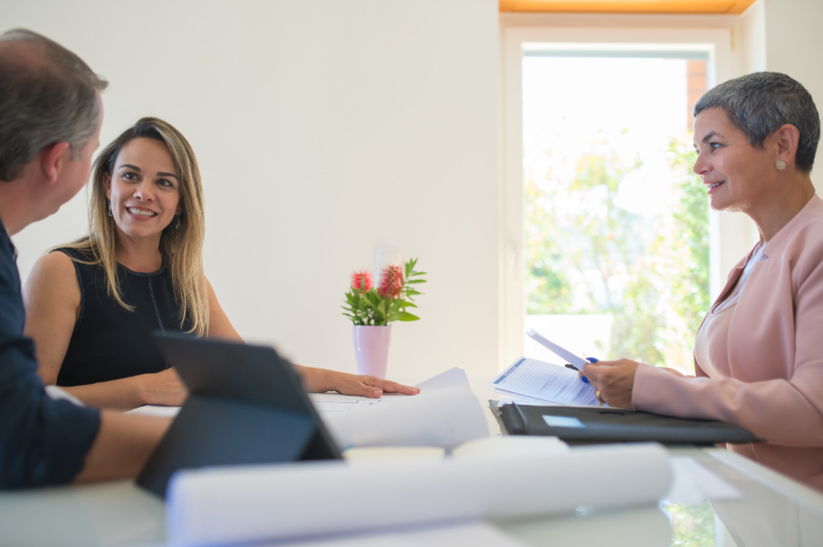 Three people in a meeting at a table, with papers, a laptop, and a vase of flowers by a bright window.