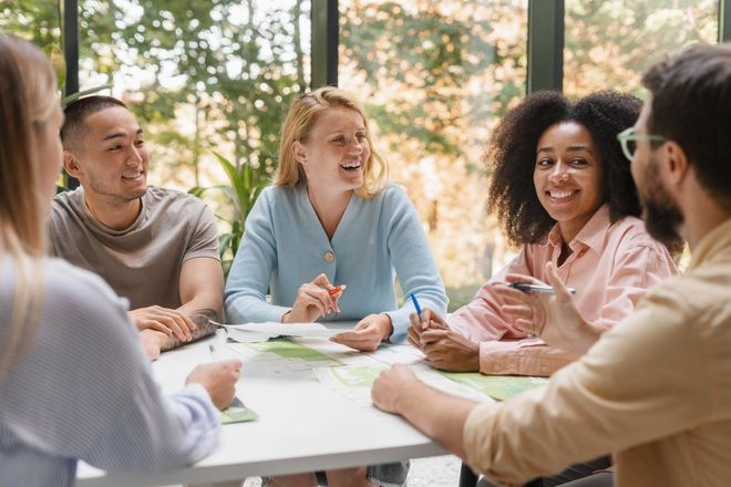 A diverse group smiles and laughs around a white table in a bright room, papers and pens in hand, collaborating.