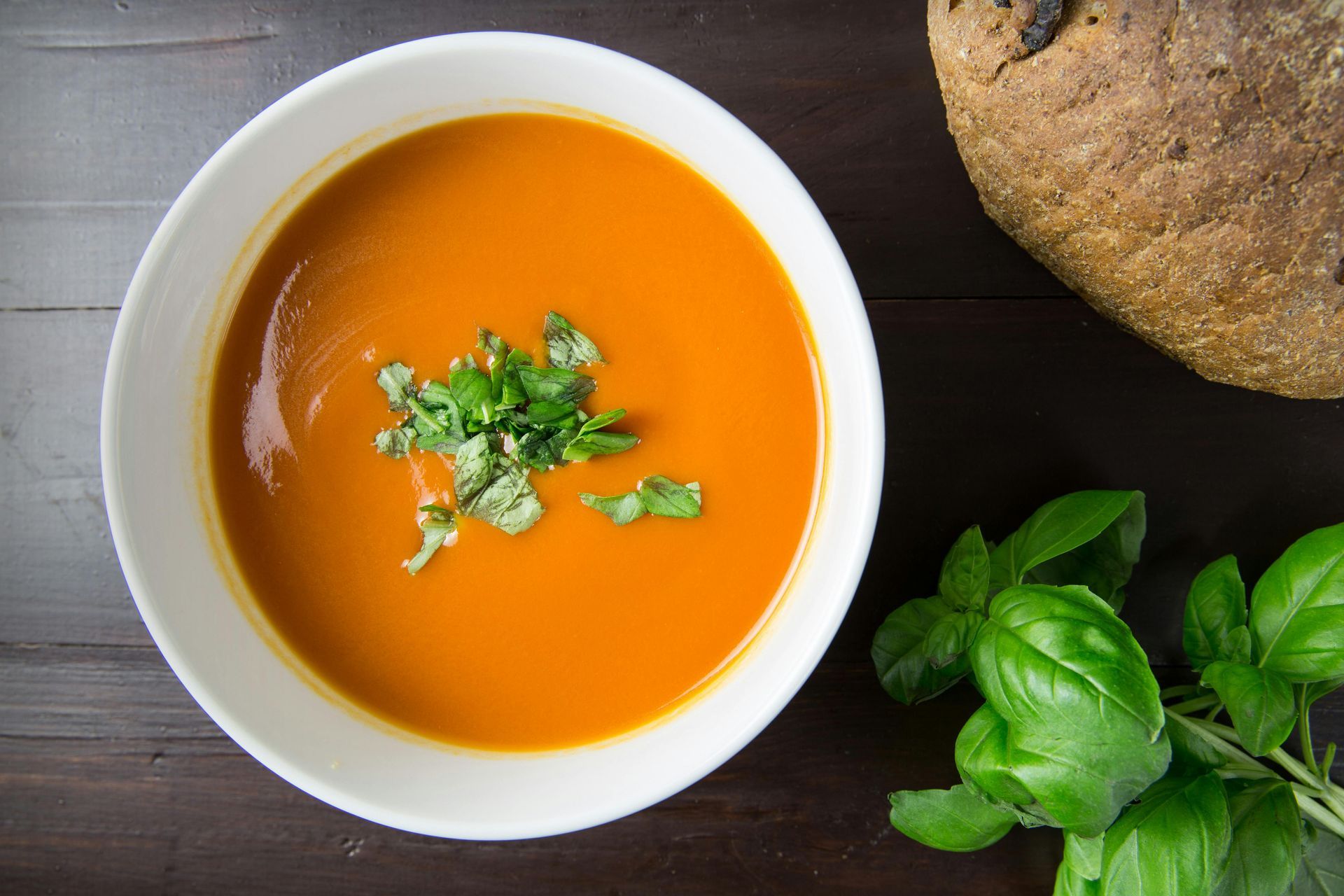 Bowl of orange soup garnished with herbs on a dark table, beside basil leaves and bread