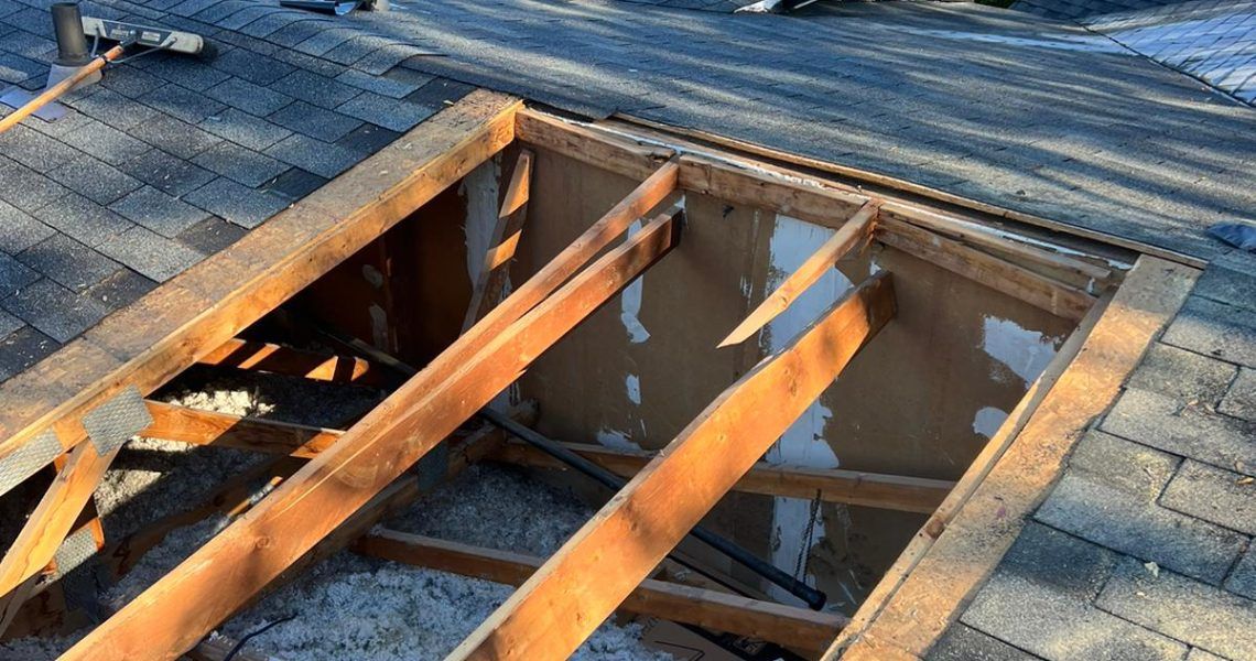 Exposed wooden roof framing around a rectangular opening in a shingled roof, with daylight below