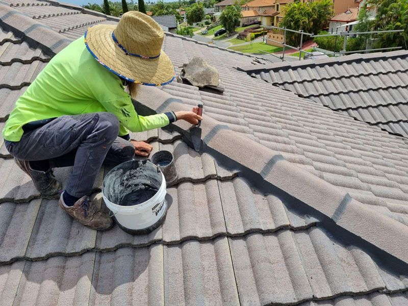 Worker painting a gray tiled roof with a paint sprayer and bucket nearby