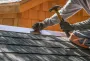 Roofers installing shingles with a hammer on a gray roof against a wooden house wall