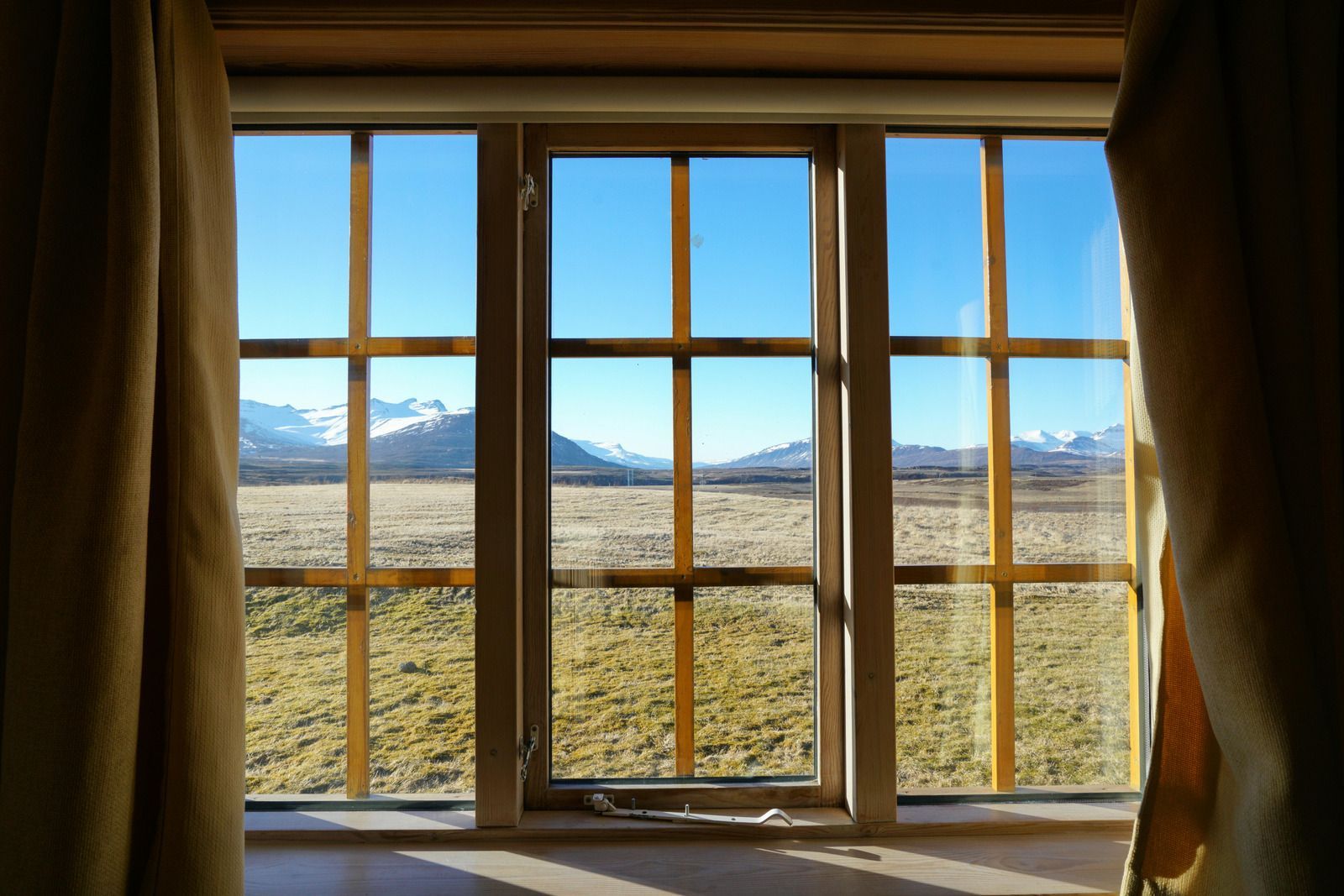 Sunlit window with wooden frames overlooking a grassy plain and snow-capped mountains under a blue sky