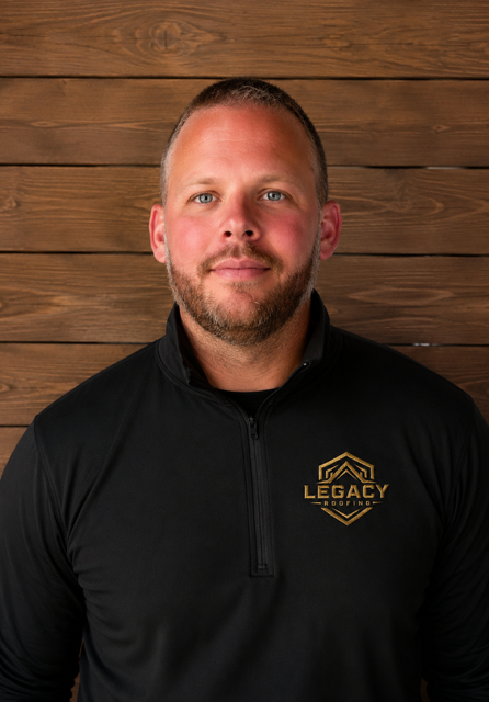 Man in black shirt with gold logo, standing against a wooden wall background.
