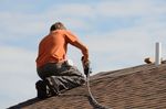 Worker in orange shirt and black pants using a nail gun on a shingled roof under a blue sky