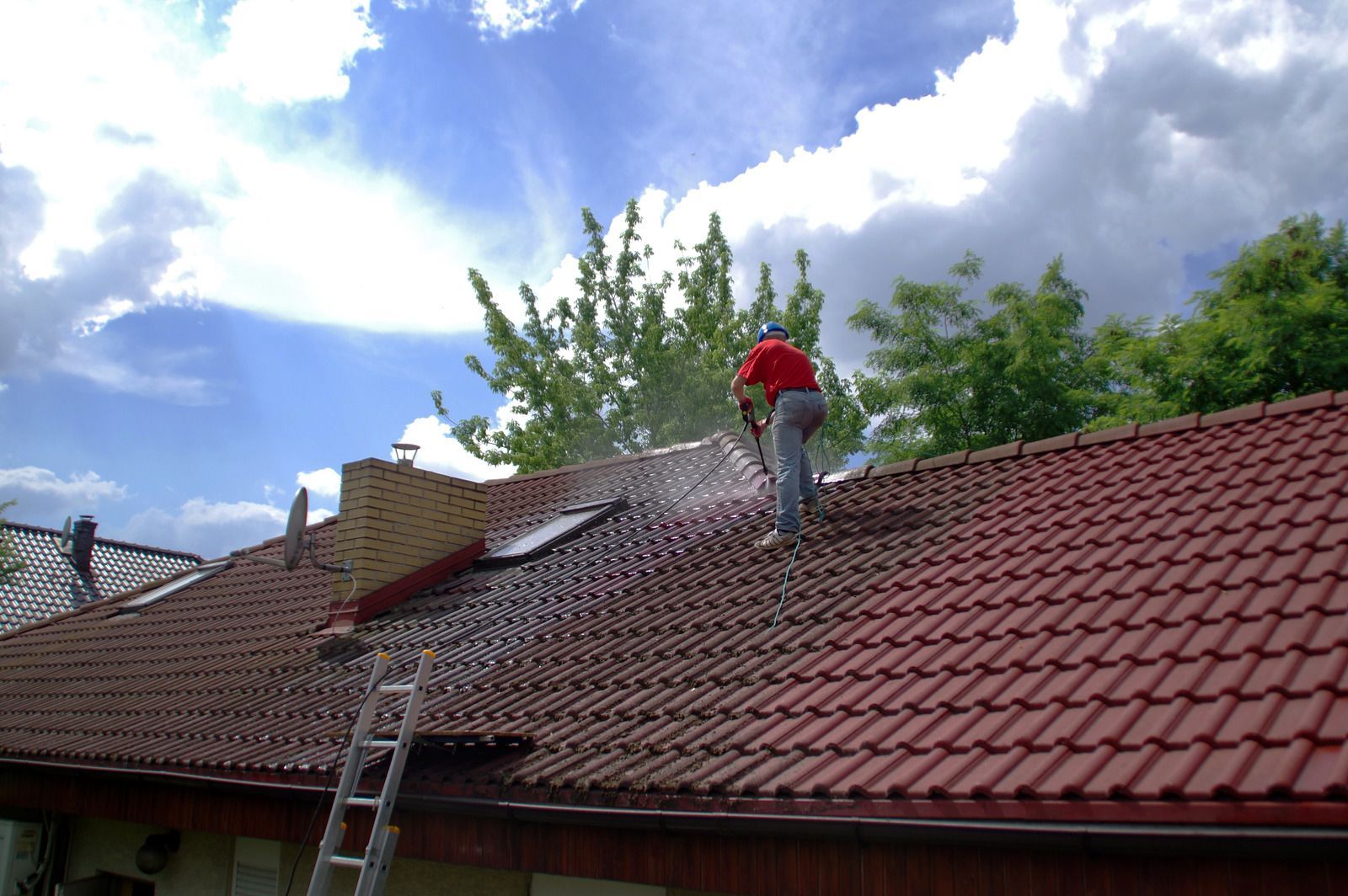 Person on a red-tiled roof near a ladder, with trees and cloudy sky in the background