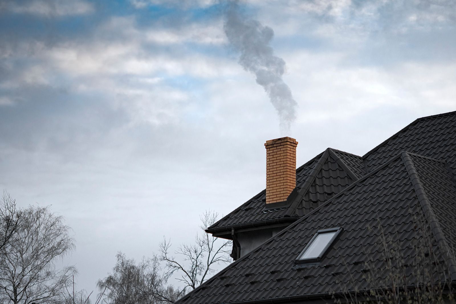 Smoke rising from a brick chimney on a dark roof under a cloudy sky.