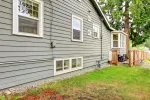 Side of gray house with white-trimmed windows, small deck, and green yard with trees nearby.