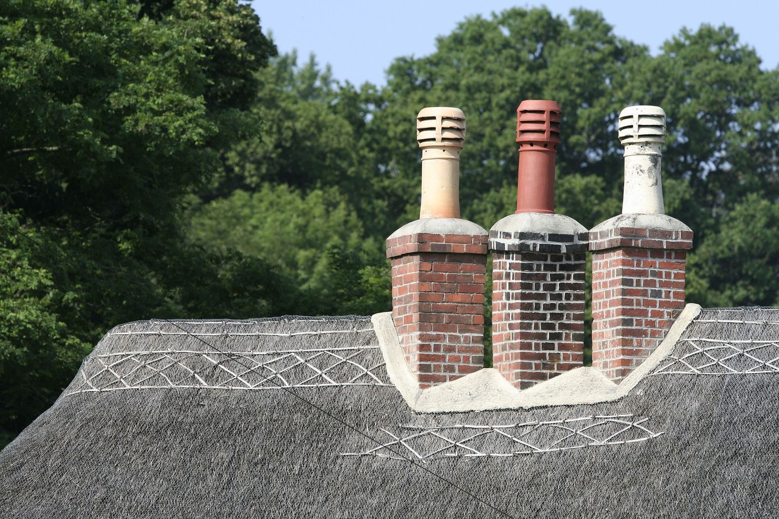 Three brick chimney pots rising above a gray shingled roof with green trees in the background