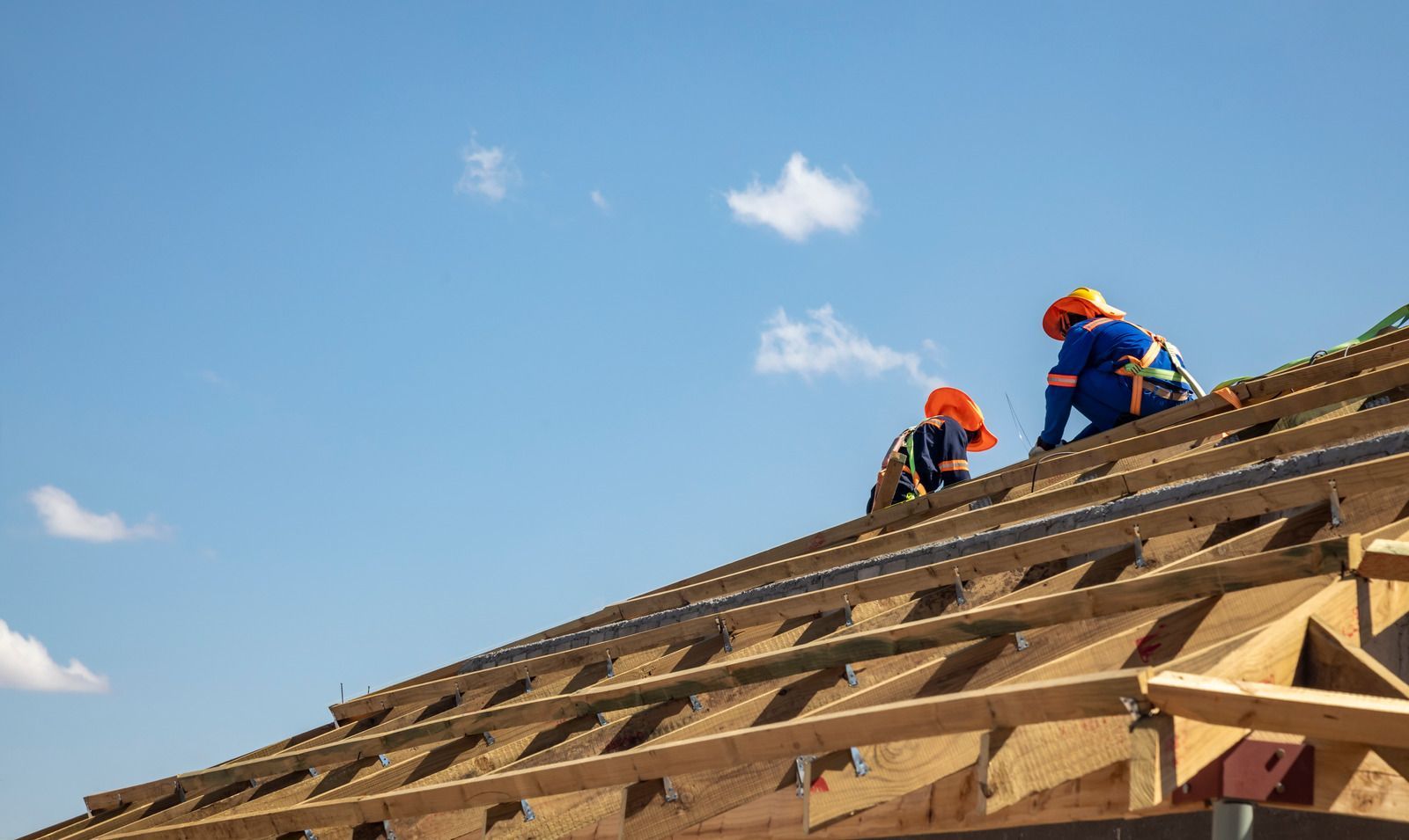 Two workers in orange gear installing roofing on a sloped wooden roof under a blue sky
