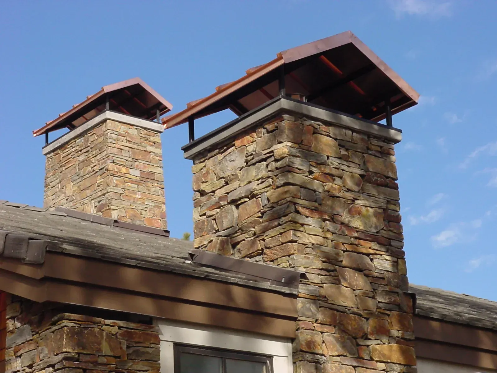 Stone chimneys with metal caps on a rustic roof against a blue sky